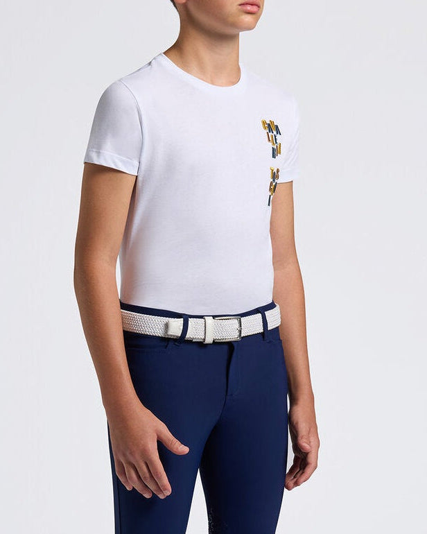 A young male model wearing a white short-sleeve t-shirt with a small embroidered crest on the left chest, navy blue equestrian riding breeches with a silver belt, and black riding boots, standing against a light gray studio background.