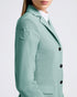 A close-up side view of a model wearing a mint green textured blazer with a tonal embroidered logo patch on the sleeve, paired with a white top underneath, against a light gray studio background.