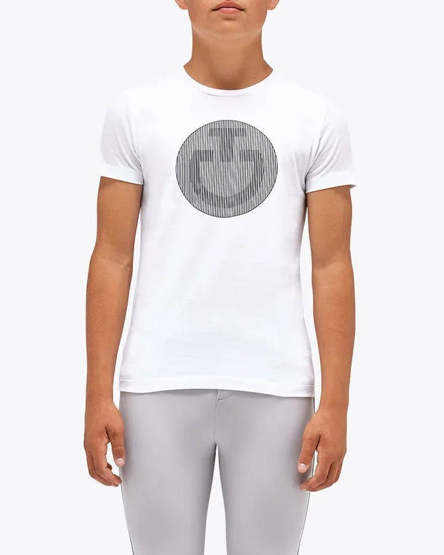 A young male model standing against a light gray studio background, wearing a white short-sleeve T-shirt with a gray circular chest graphic, light gray fitted pants, and tall black riding boots.