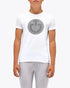 A young male model standing against a light gray studio background, wearing a white short-sleeve T-shirt with a gray circular chest graphic, light gray fitted pants, and tall black riding boots.