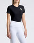 A woman modeling equestrian apparel, featuring a black short-sleeve shirt with a small white logo on the chest, high-waisted white riding breeches, and black riding boots, set against a plain light gray studio background.