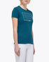A woman modeling a teal short-sleeve equestrian t-shirt with a white geometric patterned chest graphic, paired with white riding breeches and black riding boots, against a plain light gray background.