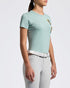 A girl stands in profile wearing a mint green short-sleeve equestrian shirt with a small embroidered crest, light gray riding breeches, a white belt, and black riding boots against a clean white studio background.