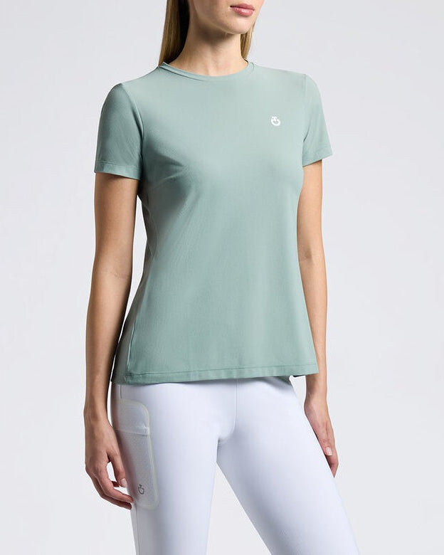 A woman modeling a sage green short-sleeve athletic top, white riding breeches, and black riding boots against a light gray studio background.